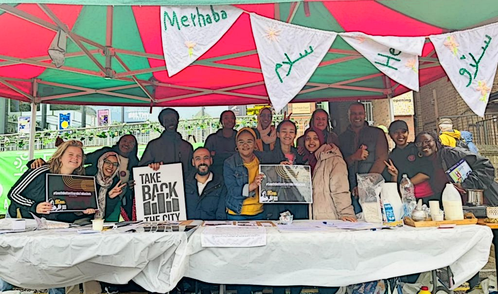 A group of people outdoors sit and stand at a market stall behind a clothed table smiling and holding "Take Back the City" and "Kind Economy" signs 