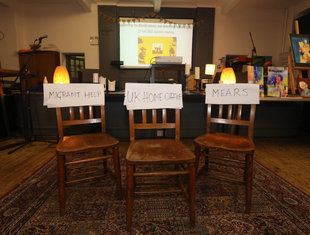 Three empty chairs with signs attached: "Migrant Help", "UK Home Office", and "Mears"