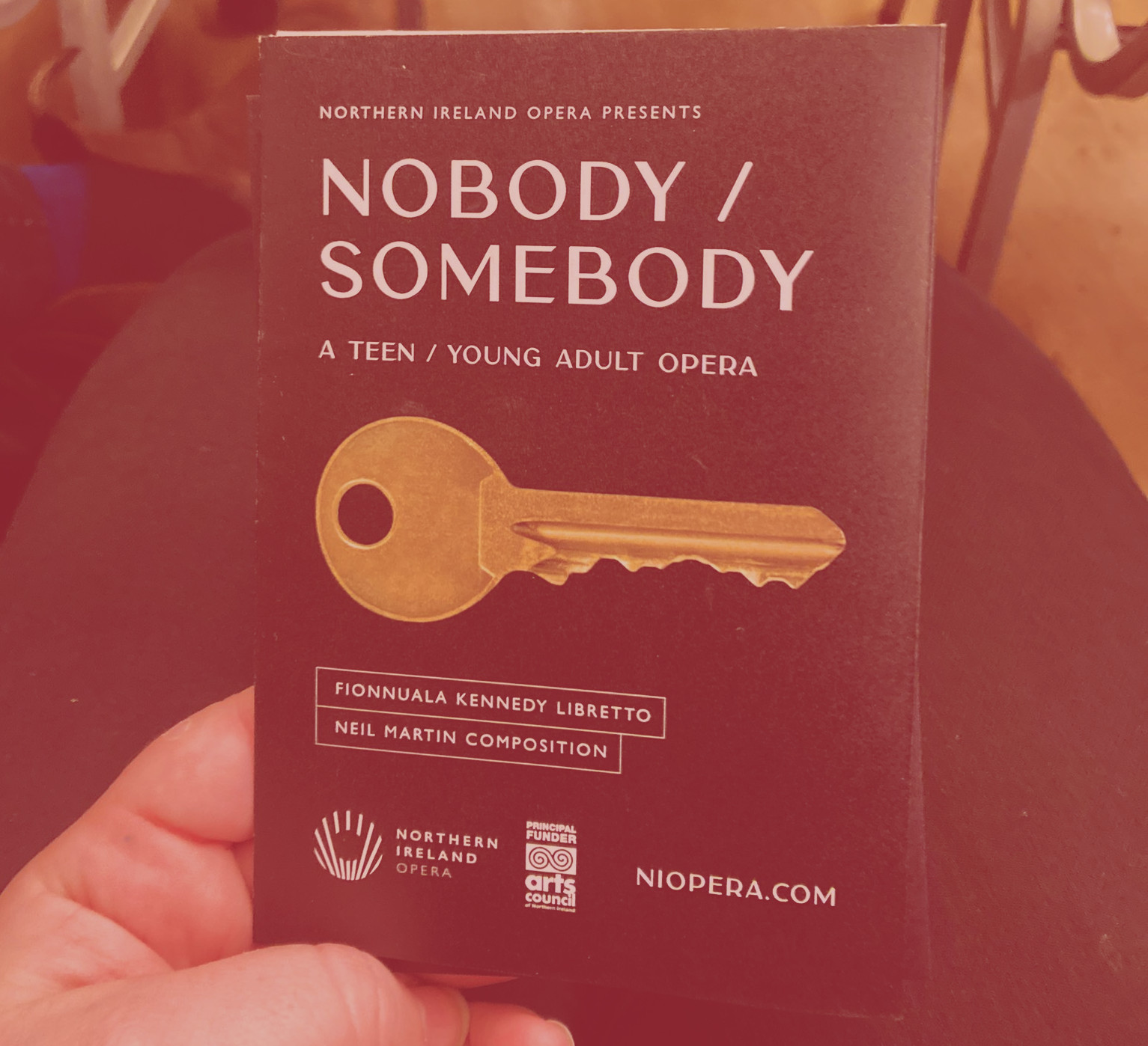 An audience member takes a photograph of his hand holding a programme for the Nobody /Somebody performace. On the cover is a golden key against a block backdrop 