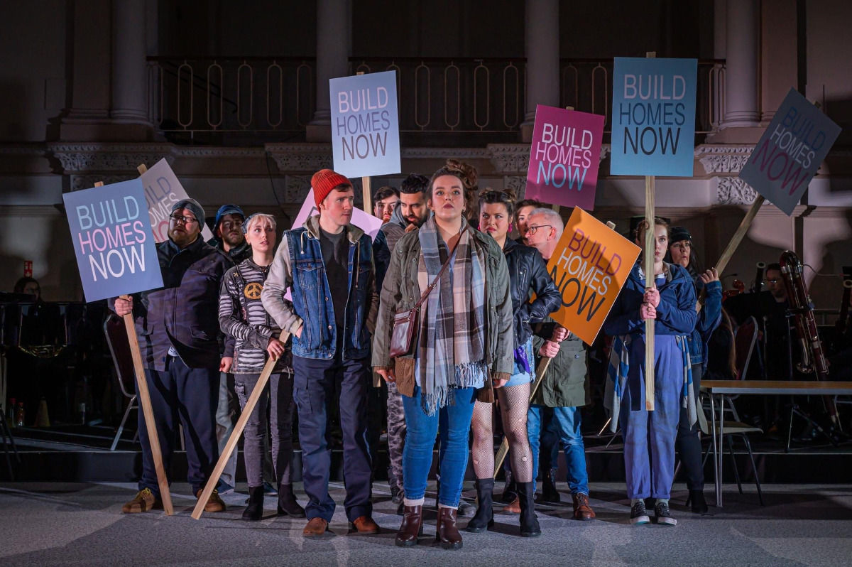 A young woman on stage stands defiantly in front of a crowd of other performers holding placards with 'Build Homes Now' written on them